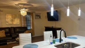 A modern kitchen with a dishwasher, a sink, and a countertop. There is a dining area with a sofa and a television in the background.