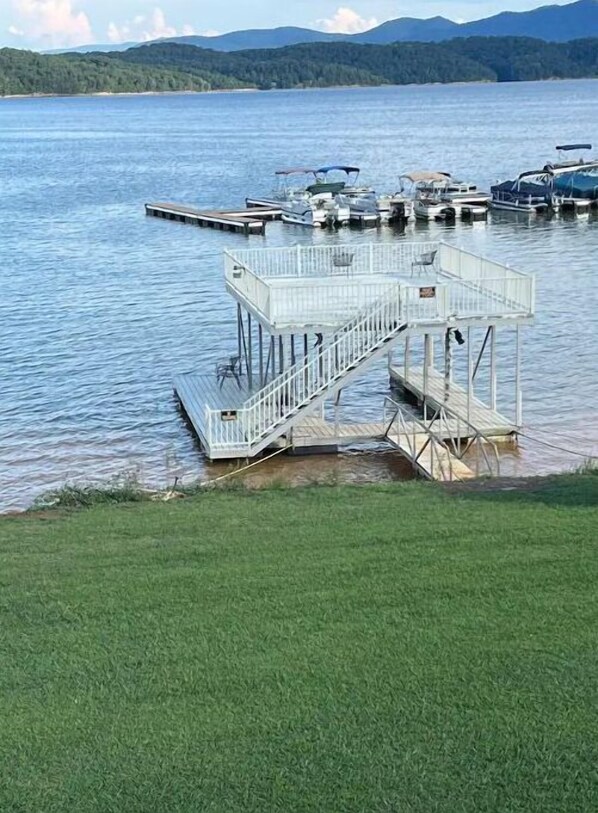 Un muelle con una pequeña estructura blanca, posiblemente un área de lanzamiento de barcos o una plataforma de observación, que se extiende sobre un lago tranquilo rodeado de colinas verdes.