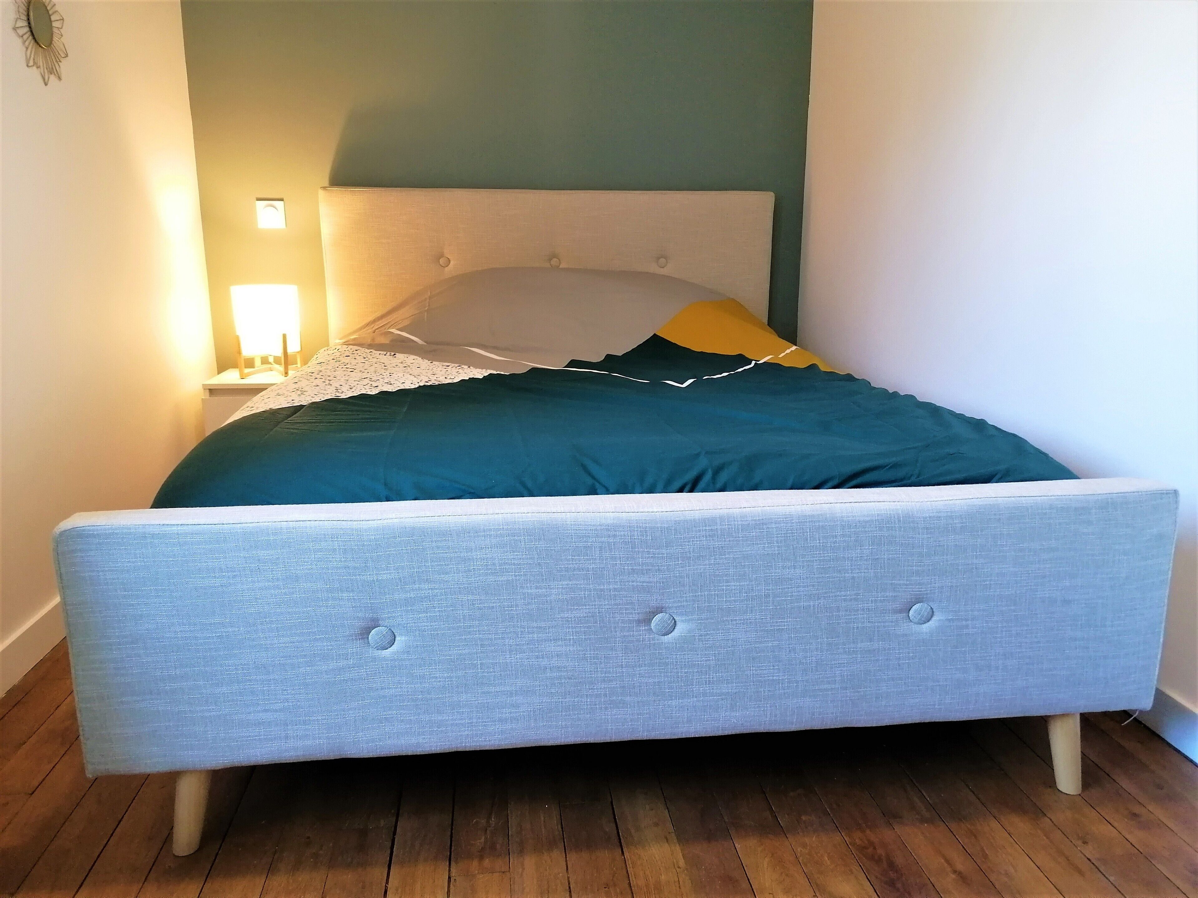A bed with a grey upholstered headboard, a teal and yellow pillow, and a white lace-trimmed pillow.