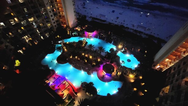 An aerial view of a hotel with a large, illuminated swimming pool surrounded by palm trees and a beach.