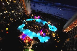 An aerial view of a hotel with a large, illuminated swimming pool surrounded by palm trees and a beach.