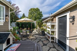 Un patio con mesa, sillas y una sombrilla grande. Hay macetas con plantas y una casita para perros.