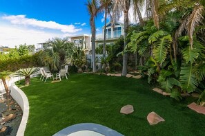 A well-maintained lawn with a few rocks, a patio area with chairs, and a large house with a balcony.