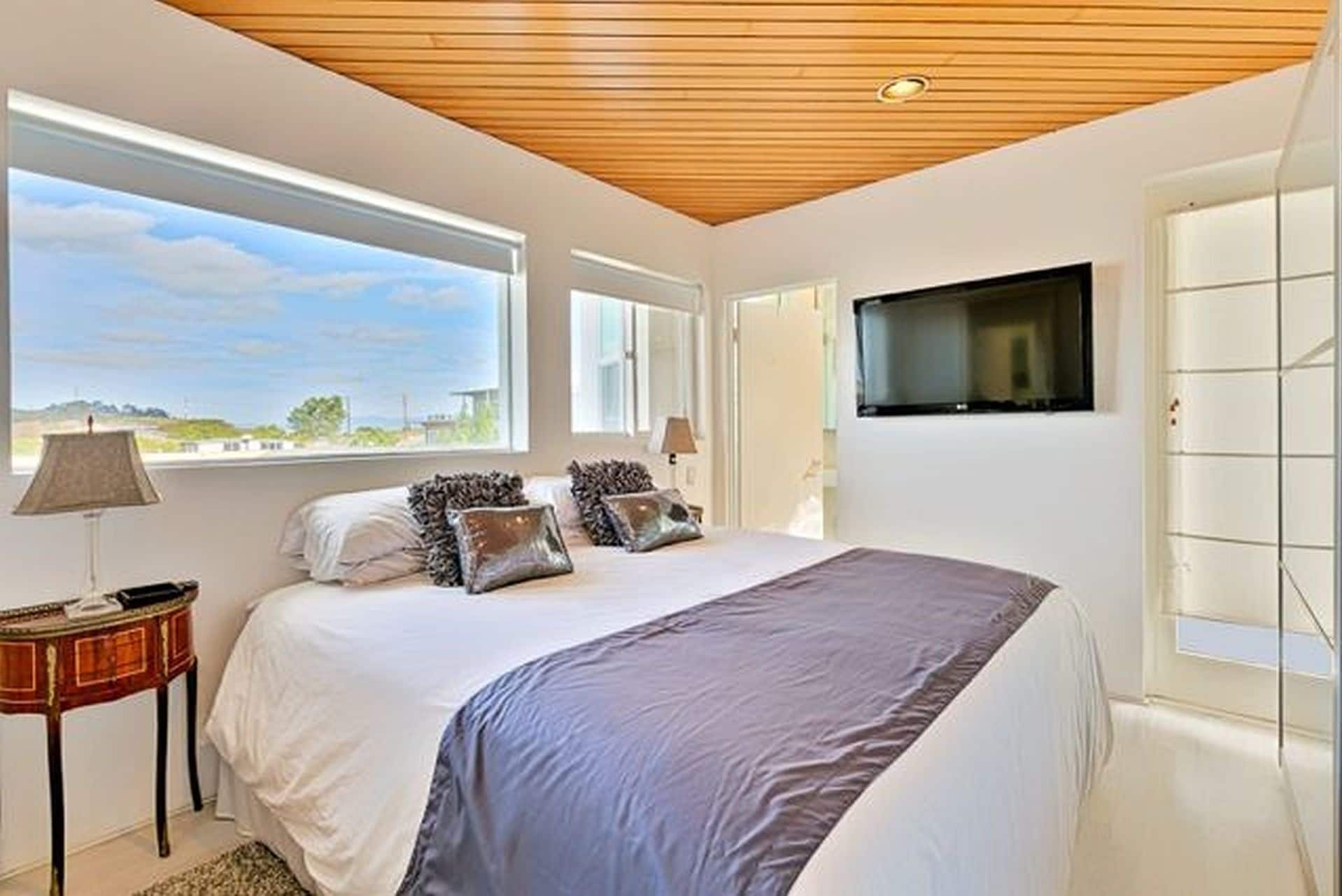 A bedroom with a large bed, a TV mounted on the wall, and a window providing a view of the outdoors.