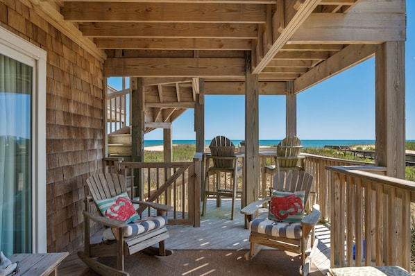 A wooden deck with two Adirondack chairs, a table, and a view of the beach.