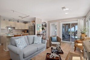 Un espace de vie moderne avec un canapé gris, des meubles en osier, une cuisine aux armoires beiges et un balcon avec vue.