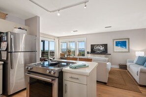 A modern kitchen with a built-in stove, refrigerator, and oven. There is a dining area with a table and chairs, and a living room with a sofa and a TV. - Sand Flee: Oceanfront condo, first level, awesome ocean views, community pool (Kill Devil Hills)