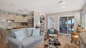 A modern living space with a grey sofa, wicker furniture, a kitchen with beige cabinets, and a balcony with a view. - Sand Flee: Oceanfront condo, first level, awesome ocean views, community pool (Kill Devil Hills)