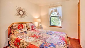 A bedroom with a wooden headboard, a floral quilt, a bedside table with a lamp, and a window with curtains. - Cozy, Secluded Davis Cabin on 60 Wooded Acres (Davis)