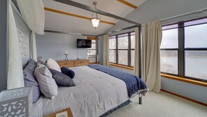 Ein Schlafzimmer mit einem großen Bett, einem Fernseher, einer Holzkommode und einem Fenster mit Blick auf ein Gewässer.