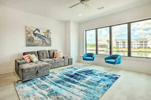 A modern living room with a grey sectional sofa, a blue rug, and a large window offering a view of buildings and water. - Lakeside Montgomery Condo Near Boat Rental! (Montgomery)