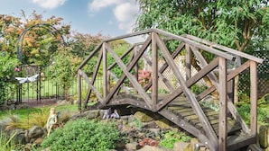 Un pont en bois enjambant un jardin de rochers, avec des statues et un kiosque en arrière-plan.