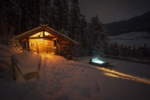 Una cabana de fusta amb llum interior, envoltada d'arbres coberts de neu i un paisatge nevós.