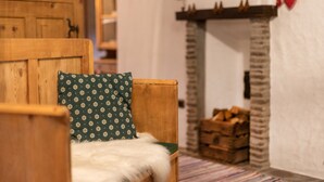 Ein gemütliches Zimmer mit einem Holzstuhl, einem grün gemusterten Kissen, einer weißen, flauschigen Decke, einem Kamin und einem Holzstapel.