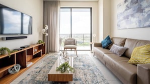 A modern living room with a flat-screen TV, wooden entertainment unit, a beige sofa, a coffee table, and a large window with a view. - Luxury Condo - 3B/3B with Amazing Skyline View (Doral)