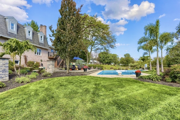 A residential backyard with a swimming pool, a lawn, and a house with a brick exterior and a sloped roof.