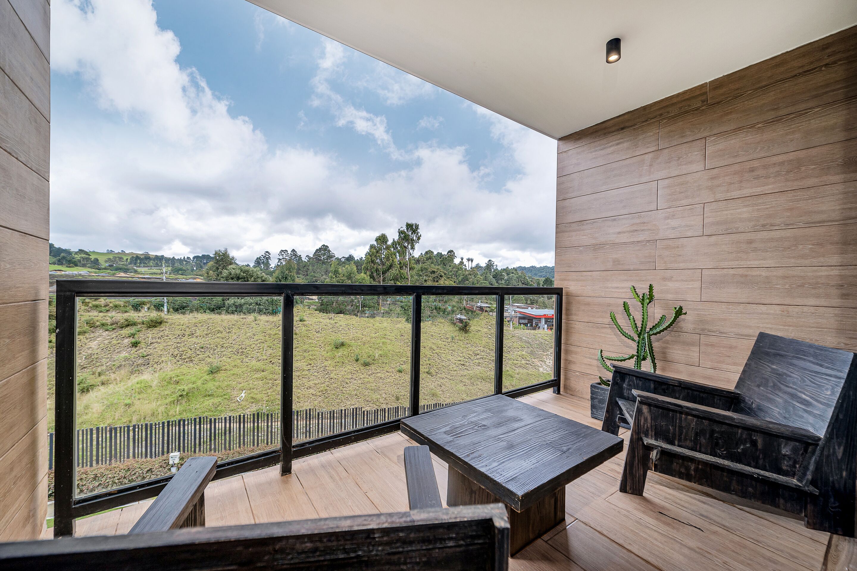 Ein Balkon mit Holzboden, einem Tisch und einem Stuhl. Im Hintergrund ist eine Wiese und Bäume zu sehen.