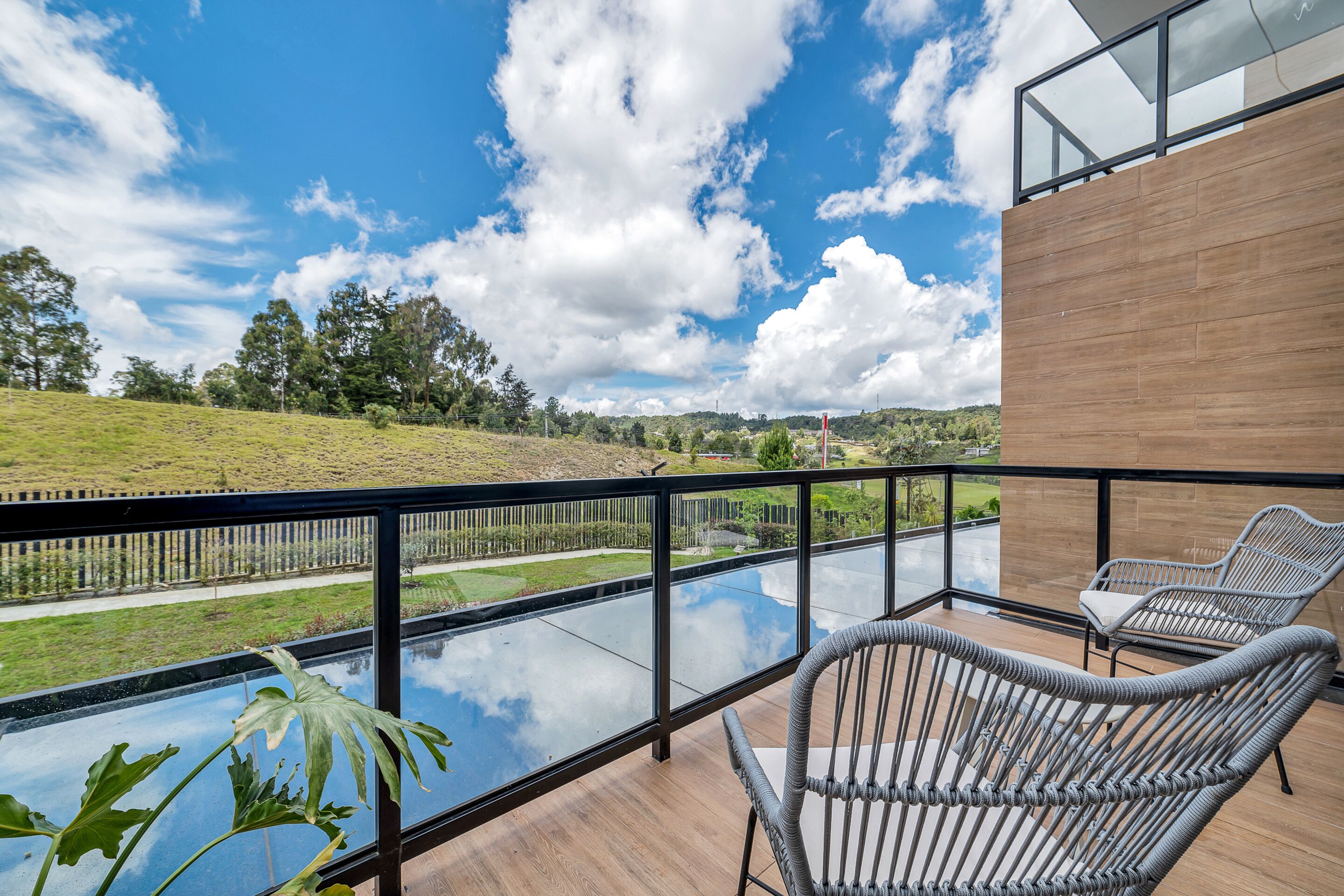 Una terraza moderna al aire libre con sillas de mimbre, baranda de vidrio y vistas a una piscina y paisaje verde.