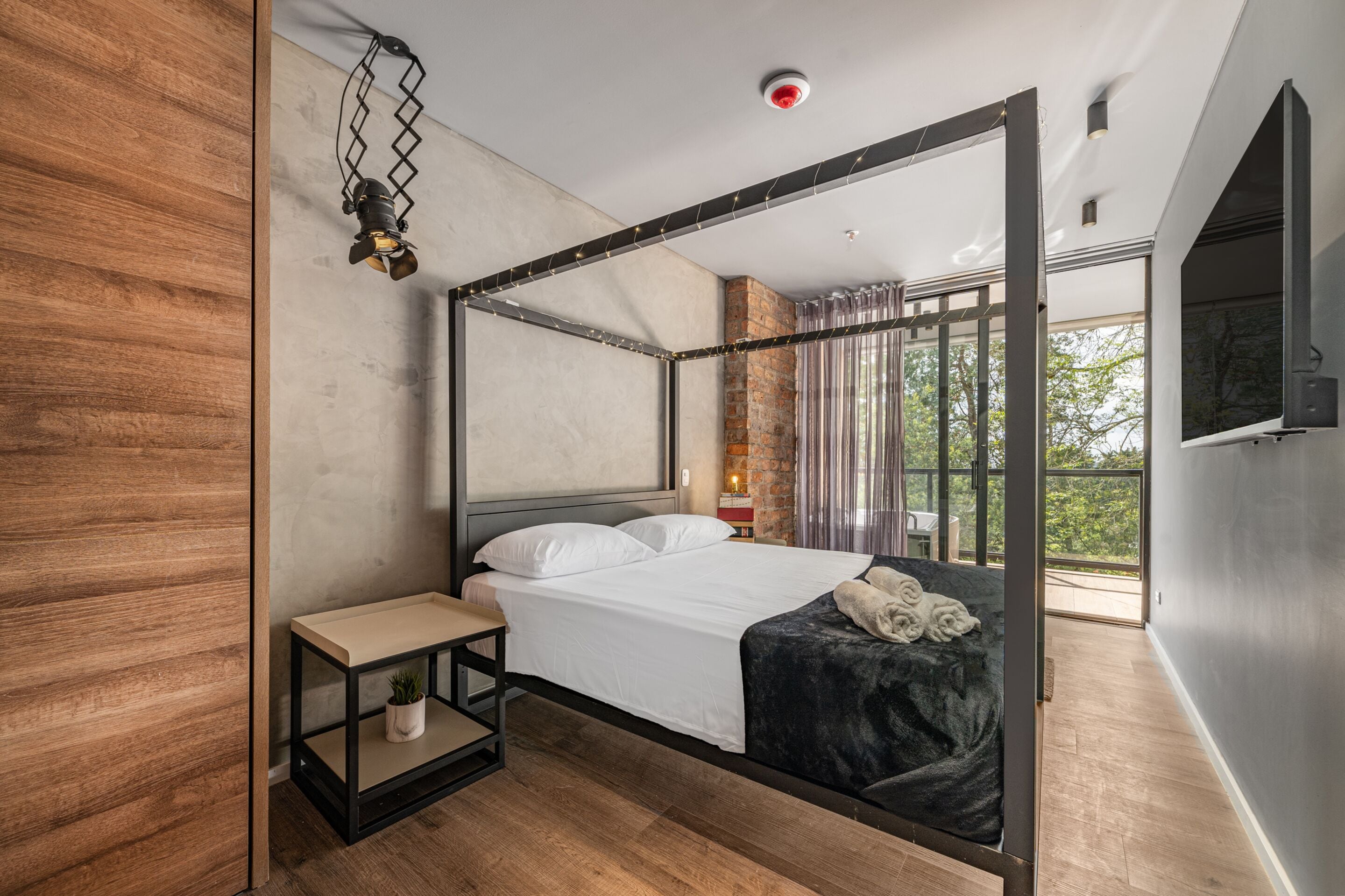 Un dormitorio moderno con una cama con dosel, una mesita de noche pequeña y un buró, y un gran ventanal con vistas al exterior.