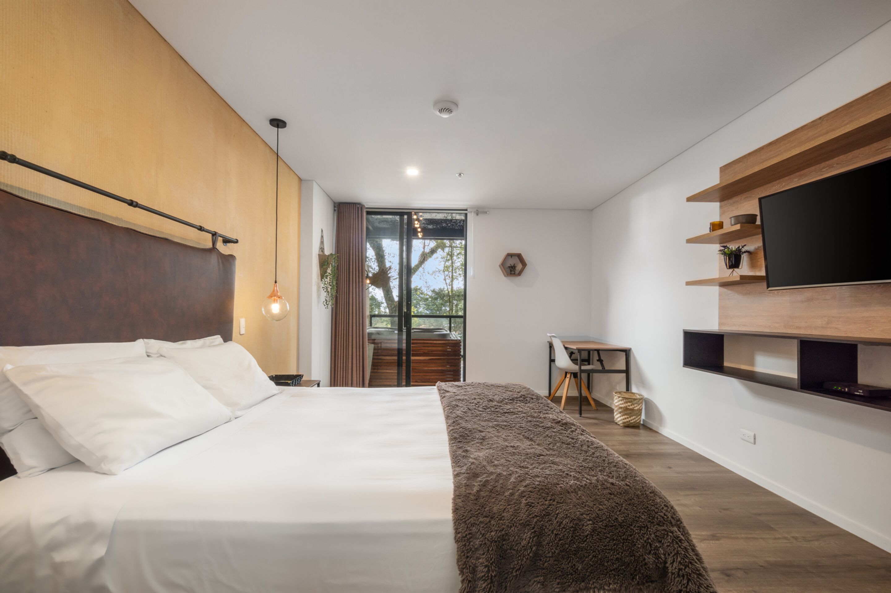 A modern bedroom with a large bed, a TV mounted on the wall, and a small desk area.