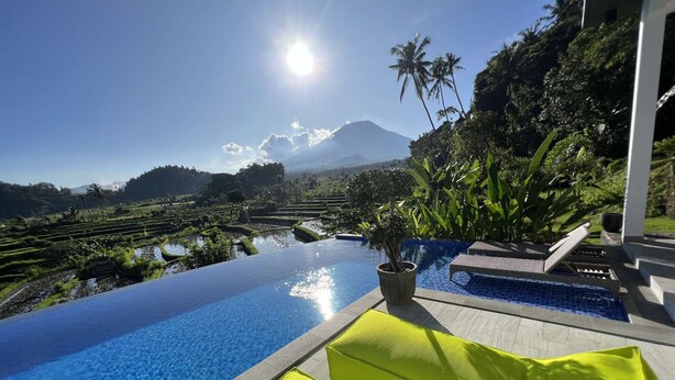 Une piscine à débordement offrant une vue sur des champs en terrasses et une montagne, le tout entouré de verdure et d’un bâtiment blanc.