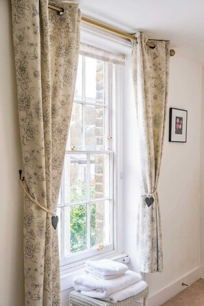 A room with a window, floral curtains, a framed picture on the wall, and a stack of white towels.