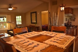 A dining area with a wooden table, four placemats with animal prints, chairs, a ceiling fan, a window with blinds, and a kitchen area in the background.