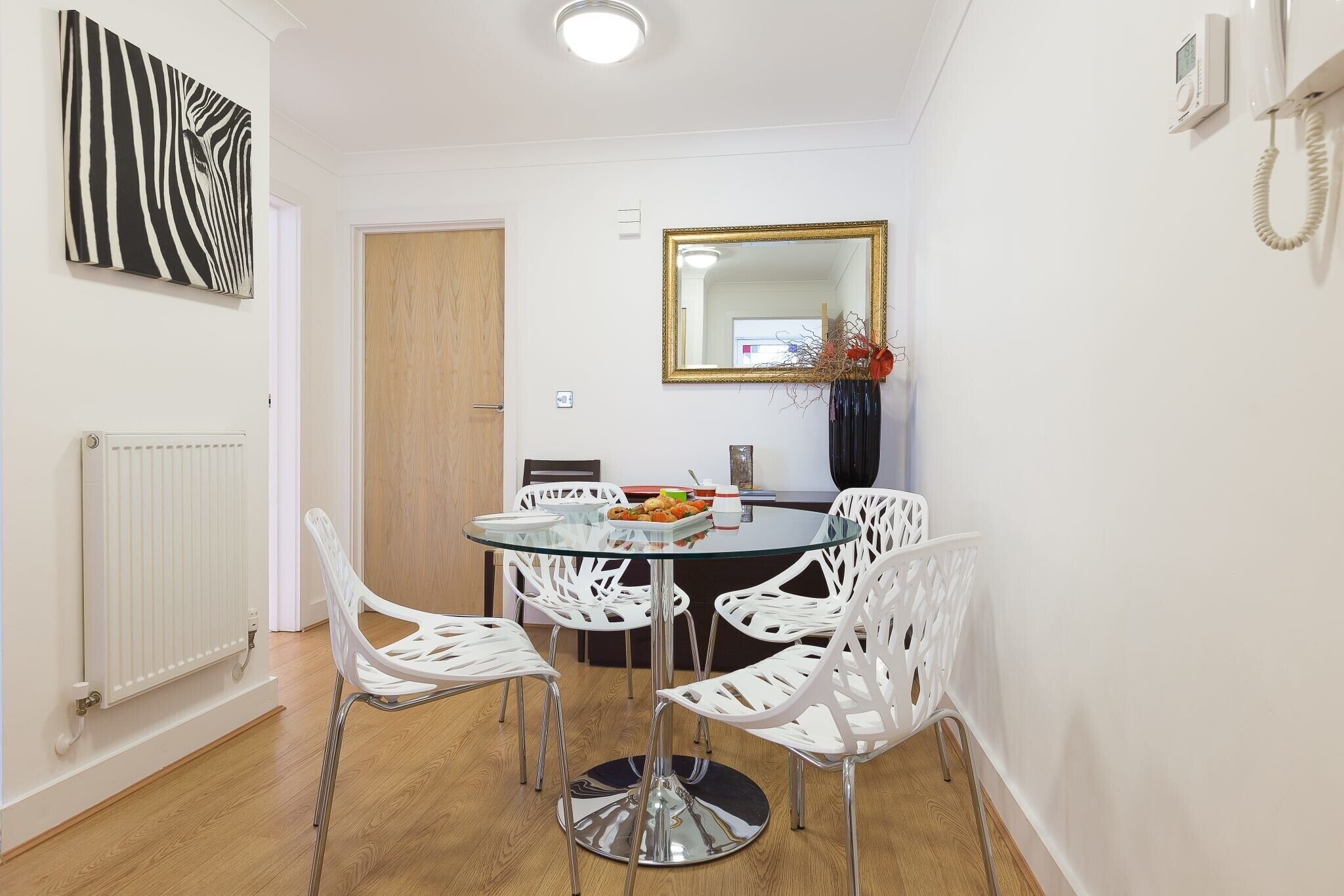 A dining area with a round glass table, white chairs, a mirror, and a radiator.