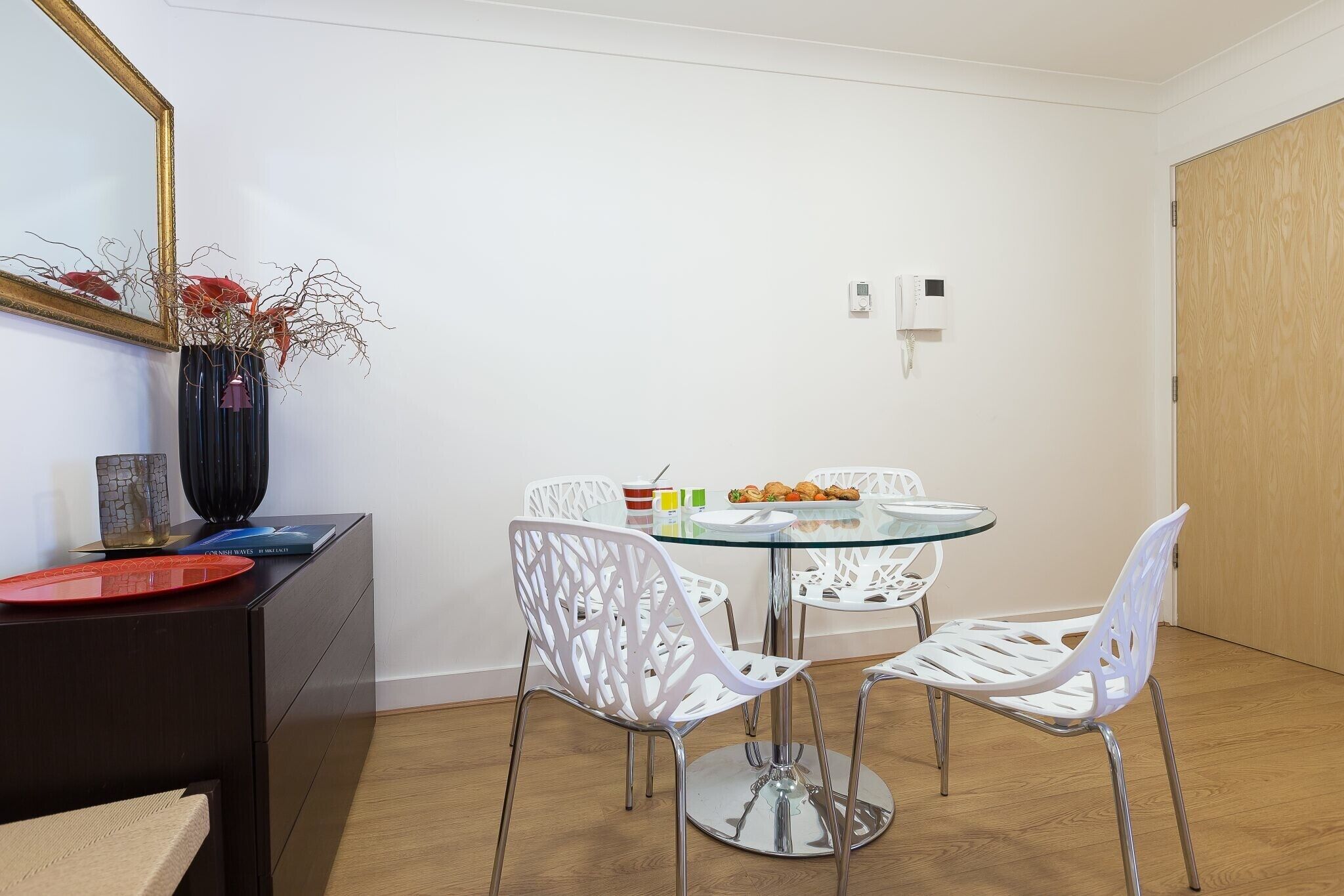 A dining area with a glass table, white chairs, and a vase with dried flowers on a dark wooden sideboard.