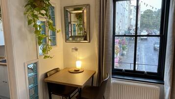 Um quarto pequeno com uma mesa e duas cadeiras, uma janela com vista para a rua, uma planta pendente e um espelho na parede.