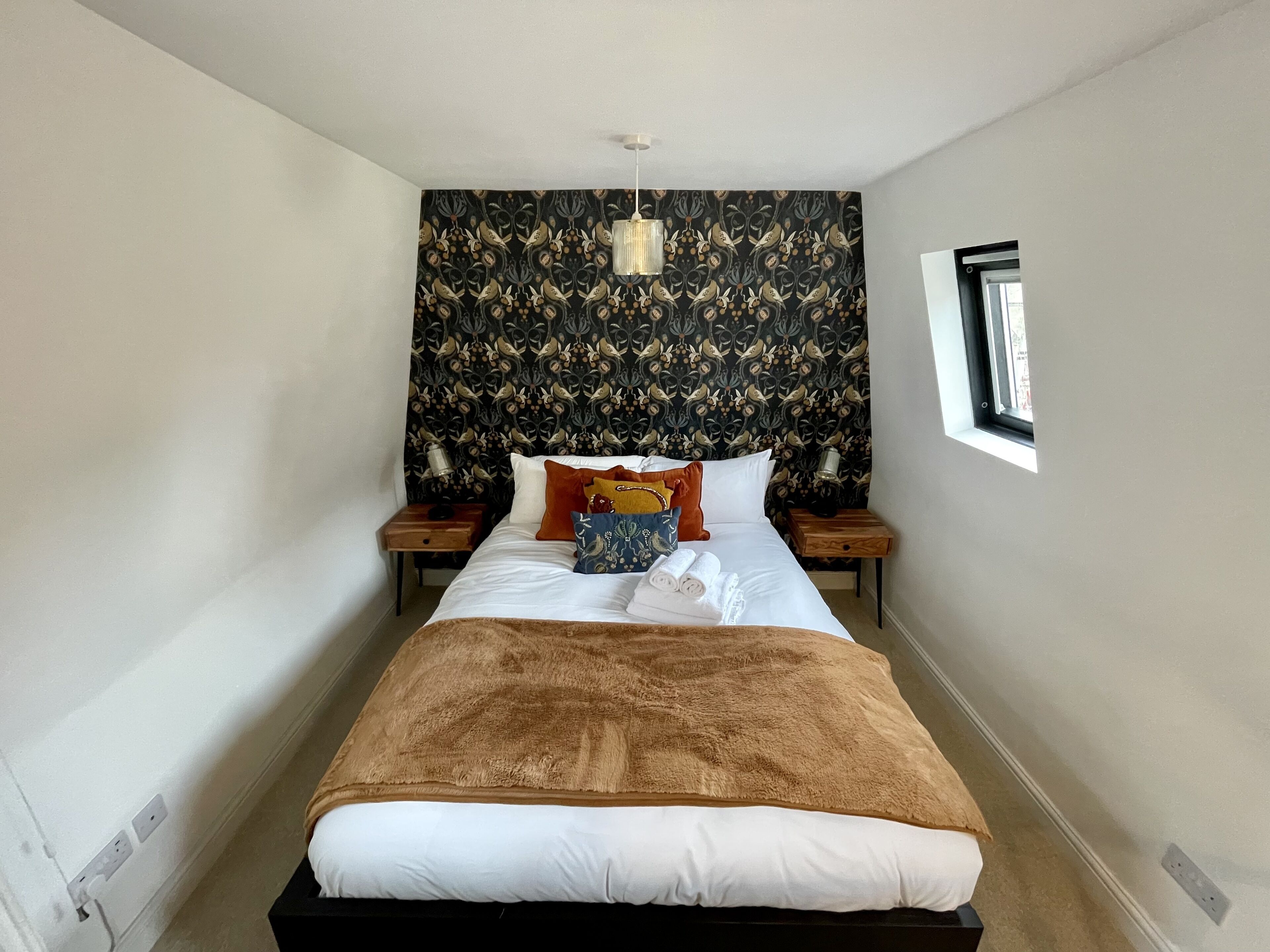 A neatly made bed with a brown blanket, white pillows, and a patterned headboard. There are two bedside tables with lamps, and a window on the right side.