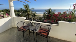 A balcony with a table and chairs, overlooking a beach with palm trees and bougainvillea flowers. - Beachfront villa is settled in Playa Agua Blanca 25 min from Puerto Escondido.  (Santa Maria Tonameca)
