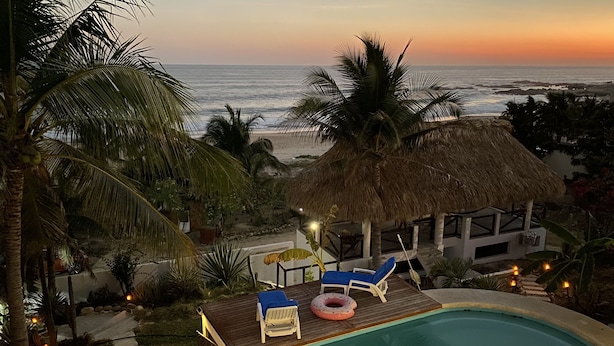 A pool area with lounge chairs and a thatched-roof structure, overlooking a beach and ocean at sunset. - Beachfront villa is settled in Playa Agua Blanca 25 min from Puerto Escondido.  (Santa Maria Tonameca)