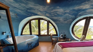A bedroom with a large bathtub, a bed, a chair, and two windows offering a view of palm trees. - Beachfront villa is settled in Playa Agua Blanca 25 min from Puerto Escondido.  (Santa Maria Tonameca)