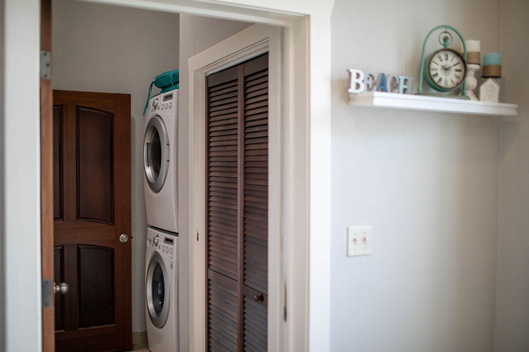 Une buanderie avec des lave-linge et des sèche-linge superposés, une porte en bois et une étagère sur laquelle se trouvent une horloge et des articles de toilette.