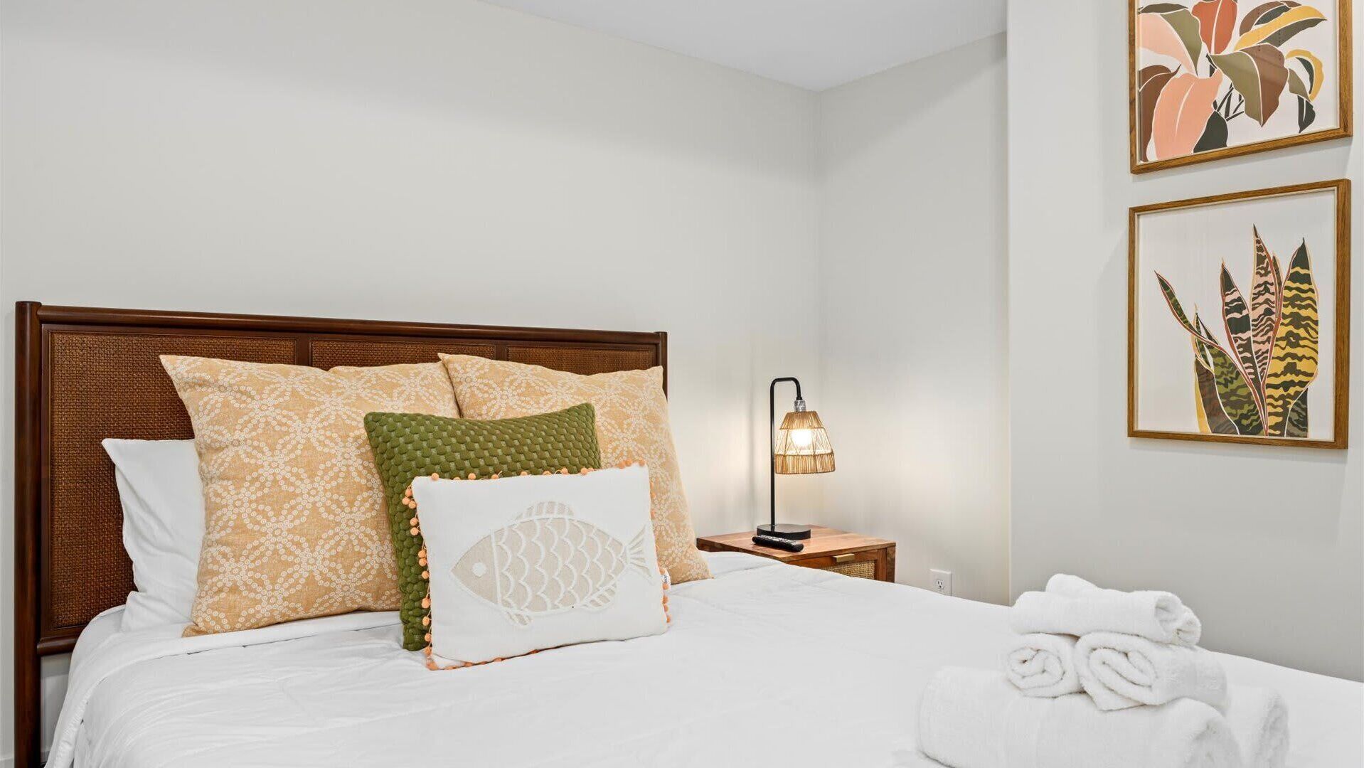 A neatly made bed with patterned pillows, a bedside lamp, and a framed plant on the wall.