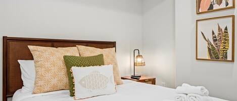 A neatly made bed with patterned pillows, a bedside lamp, and a framed plant on the wall.