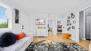 A modern living room with a white sofa, a glass coffee table, and a patterned rug. There is a hallway leading to a bedroom with a bed and a bathroom with a shower curtain. - 2BR Beach Escape - Walk to Boardwalk & Downtown OC (Ocean City)
