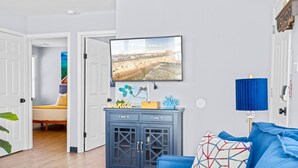 A room with a large TV mounted on the wall, a blue cabinet with drawers, a blue sofa, and a bed in the adjacent room. - Downtown OC 2BR - Walk to Beach & Boardwalk! (Ocean City)