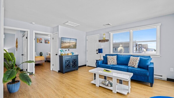A modern living room with a blue sofa, a white coffee table, and a large window with blinds. - Downtown OC 2BR - Walk to Beach & Boardwalk! (Ocean City)