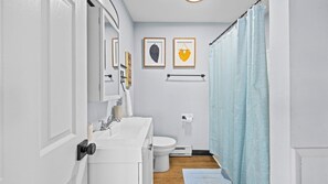 A bathroom with a white sink, mirror, and toilet. There are two framed pictures on the wall, one of a feather and one of a heart. A blue shower curtain is partially visible. - Downtown OC 2BR - Walk to Beach & Boardwalk! (Ocean City)