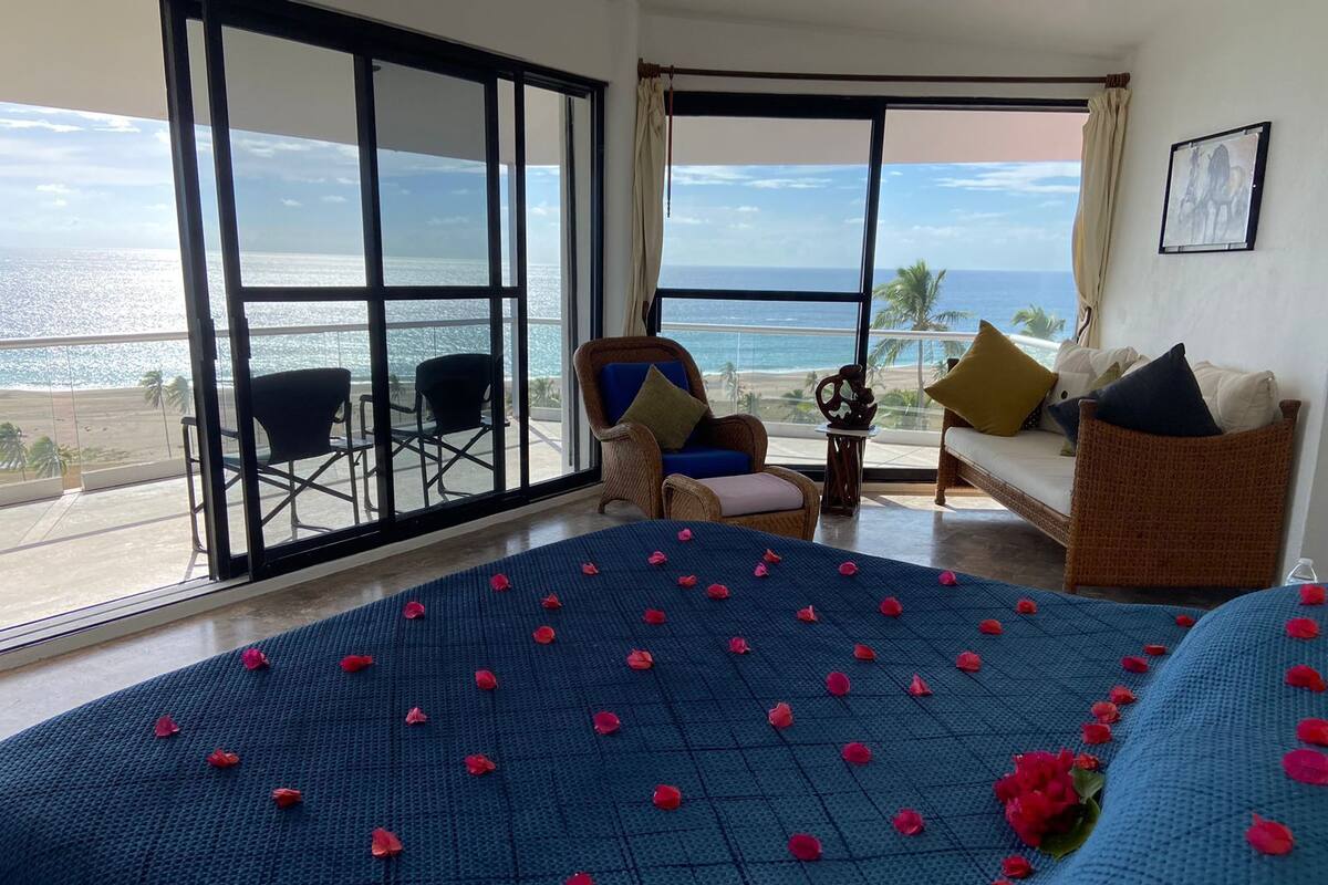 Ein Zimmer mit einem Bett, das mit Rosenblüten bedeckt ist, Meerblick und einem Balkon mit Stühlen.
