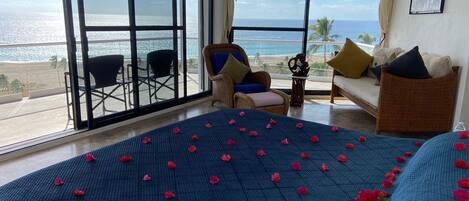 A room with a bed covered in rose petals, a view of the ocean, and a balcony with chairs.
