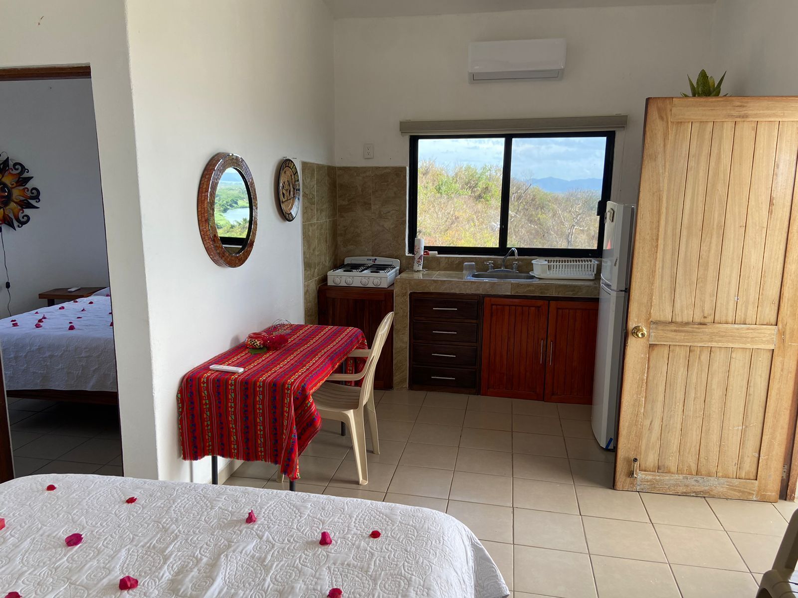 A compact hotel room with a bed, a small dining table with a red tablecloth, a kitchenette with a stove and sink, and a large window offering a view of the outdoors.
