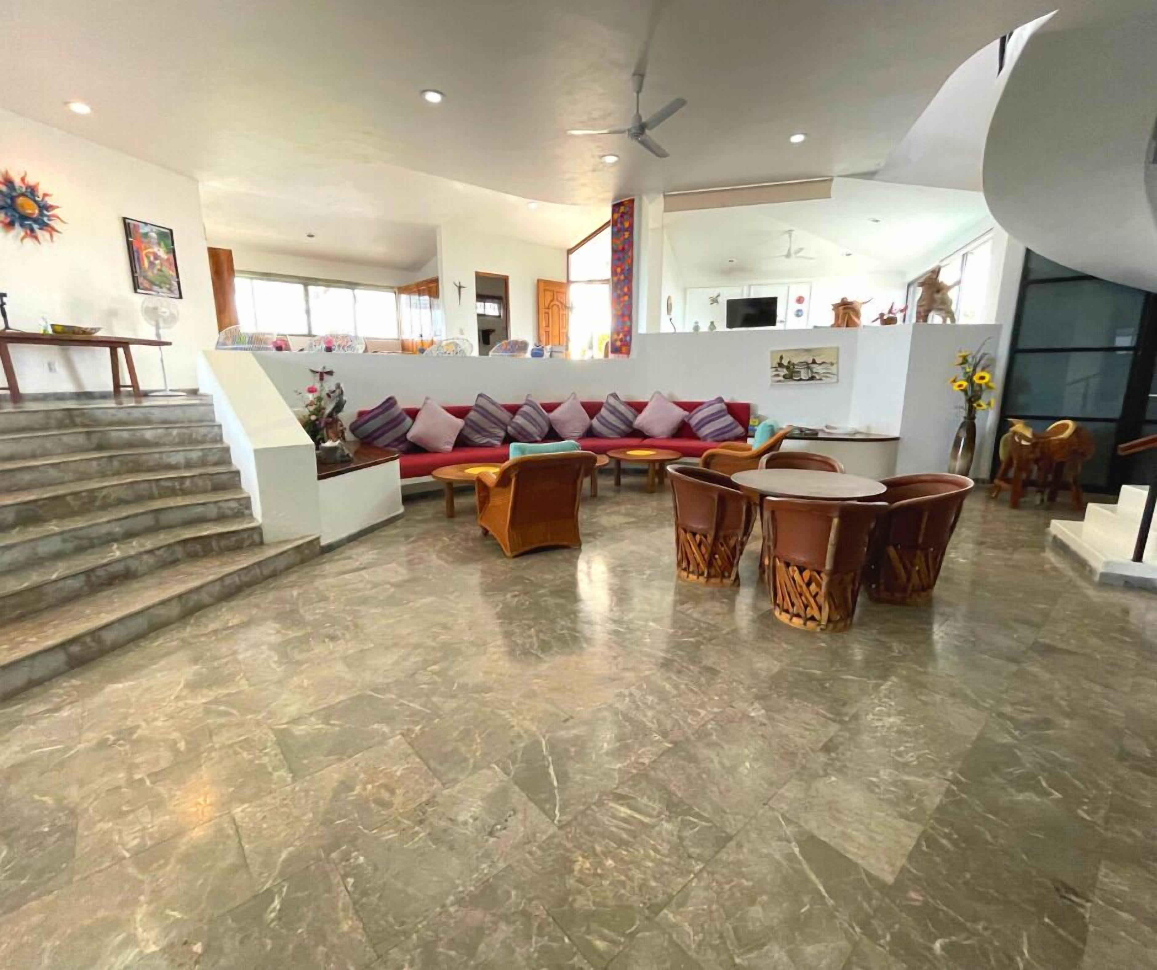 A spacious living area with a sofa set, a coffee table, and a dining table with chairs. There are stairs leading to another level, and a ceiling fan is visible.
