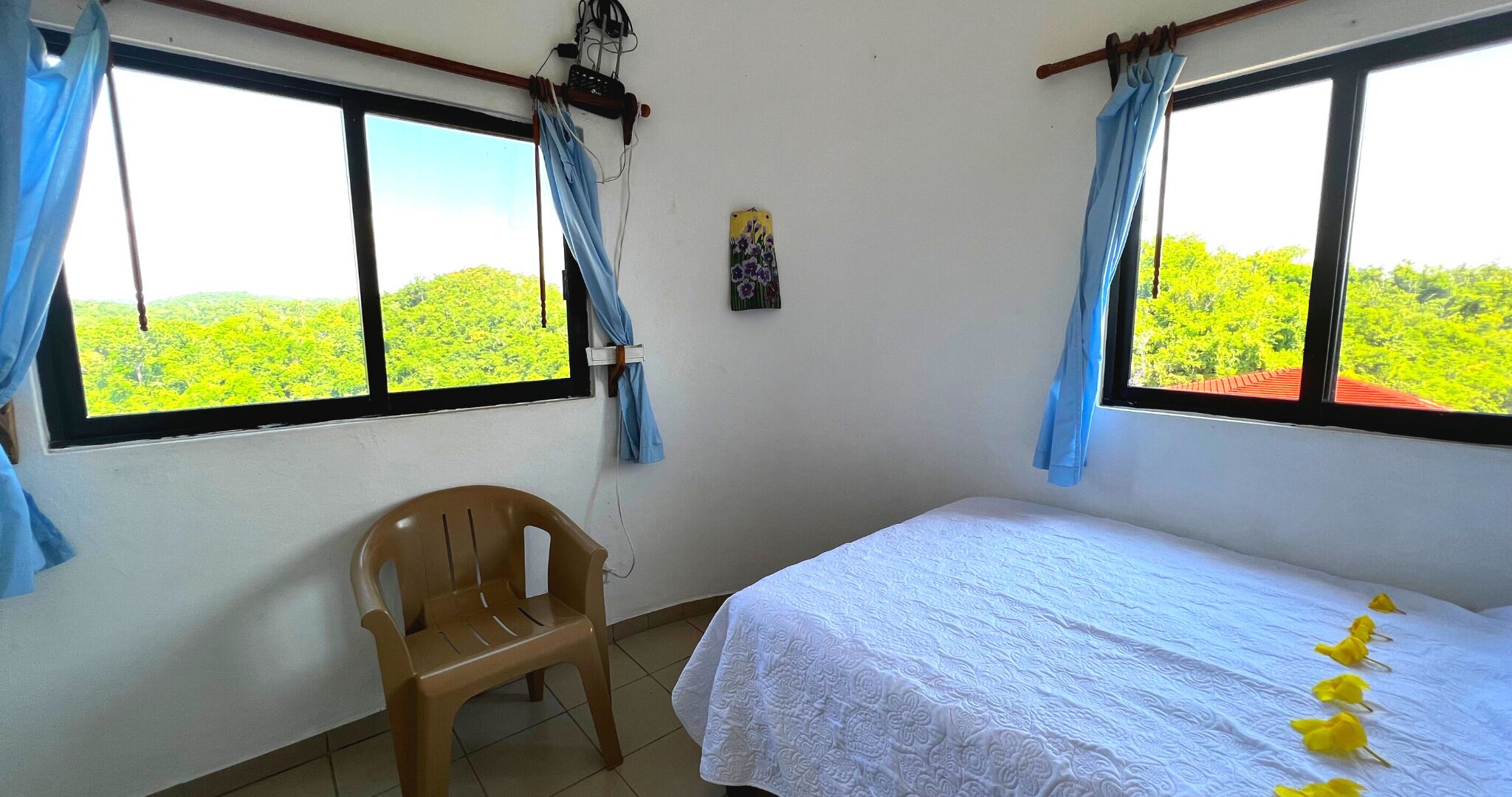 Una habitación con dos ventanas, una cama con colcha blanca, una silla de plástico y una pequeña decoración en la pared.