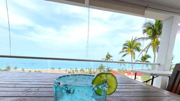 A glass of blue cocktail with a lime slice on a wooden table with a view of a beach and palm trees. - Villa Siempre Domingo (Tenacatita)