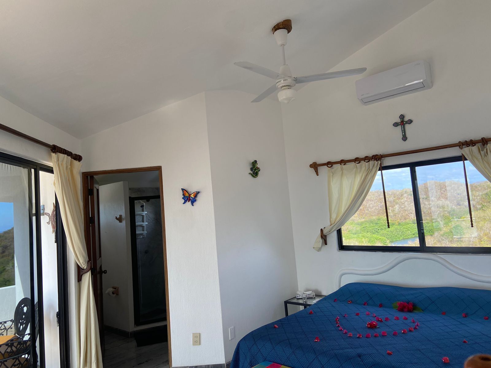 Une chambre avec un lit, un ventilateur de plafond, un climatiseur, une fenêtre avec des rideaux et une vue sur la verdure à l’extérieur.