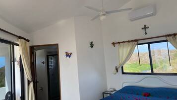 Une chambre avec un lit, un ventilateur de plafond, un climatiseur, une fenêtre avec des rideaux et une vue sur la verdure à l’extérieur.