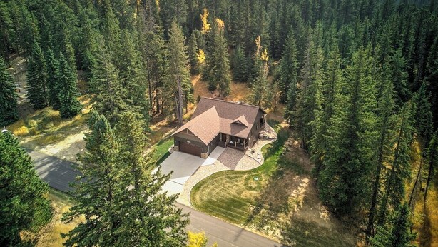 A modern house surrounded by dense forest, with a paved driveway and a well-maintained lawn.